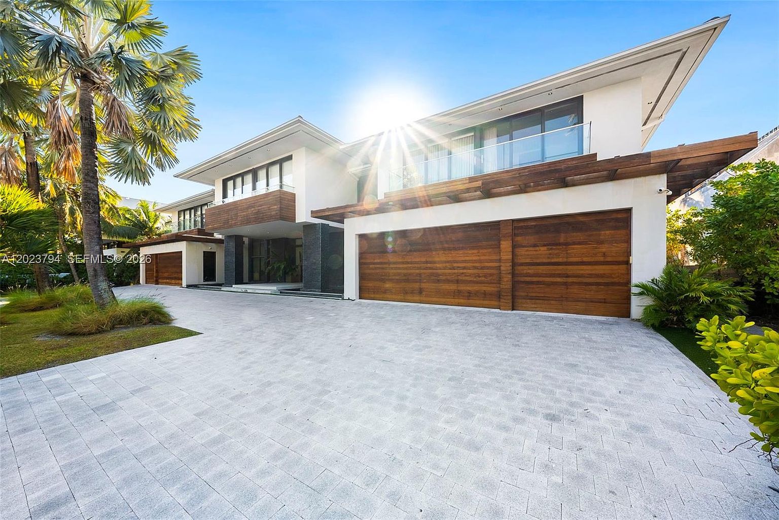 This modern luxury residence features a striking architectural design with a combination of white stucco, dark stone accents, and warm wood-paneled garage doors. The expansive paved driveway leads to a multi-car garage, while the upper level showcases a glass-railed balcony and large windows that capture the bright sunlight. The property is framed by lush tropical landscaping, including a prominent palm tree, creating a sophisticated and inviting curb appeal.