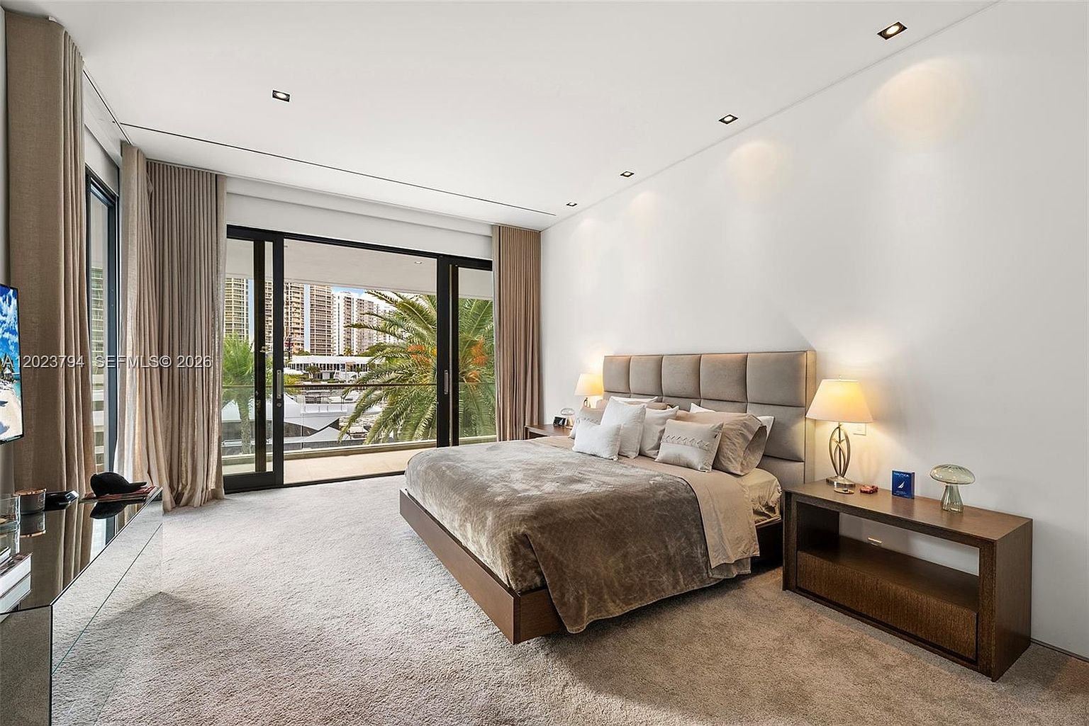 This spacious primary bedroom features a neutral color palette and a large, upholstered headboard that serves as a focal point. Floor-to-ceiling glass doors open onto a private balcony, providing ample natural light and a scenic view of the water and palm trees. The room is furnished with a low-profile bed, a dark wood side table, and a sleek console, creating a sophisticated and tranquil atmosphere.