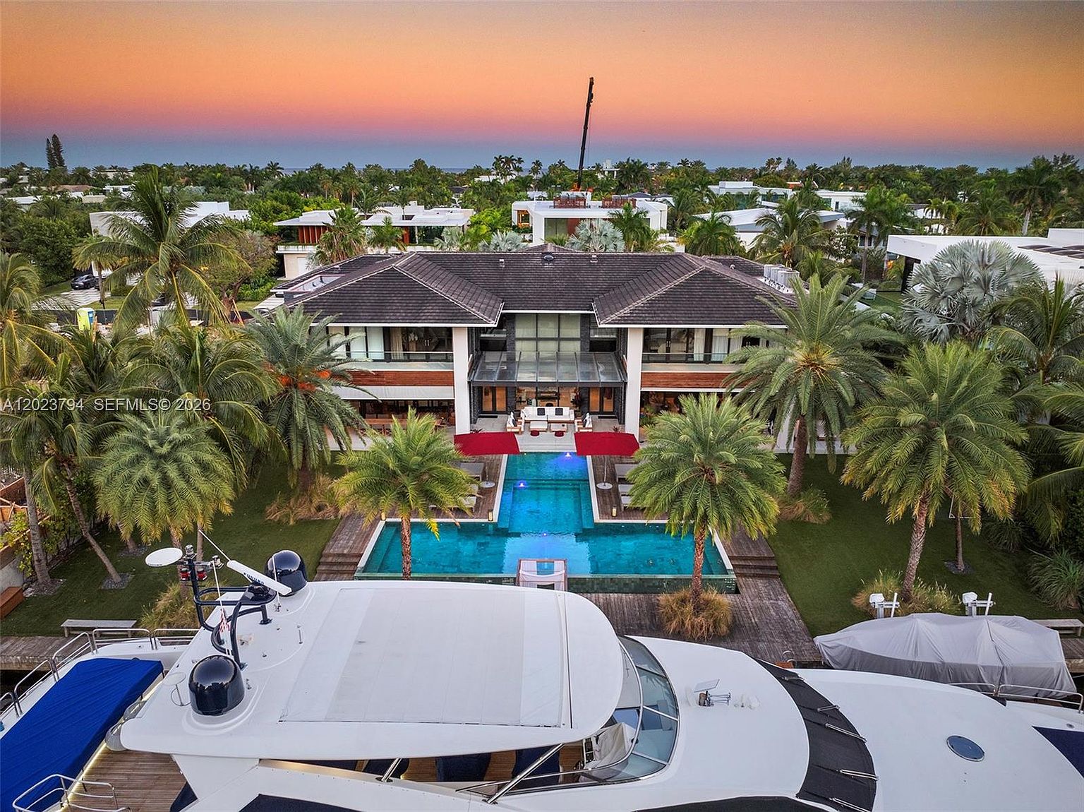 This high-angle aerial shot captures a luxurious waterfront estate at sunset, featuring a modern two-story residence with a symmetrical design and a central glass-enclosed staircase. A long, rectangular swimming pool with red umbrellas extends from the rear patio toward the water, flanked by lush palm trees and manicured landscaping. The foreground includes the top deck of a large yacht, emphasizing the property's prime location for boating and leisure.