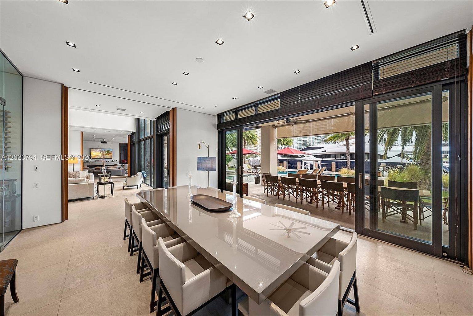 This sophisticated dining area features a long, glossy table surrounded by modern upholstered chairs, set against a backdrop of neutral tones and clean lines. Large sliding glass doors open to an expansive outdoor patio, seamlessly blending indoor and outdoor living spaces with a view of a docked yacht. The room is illuminated by recessed lighting and offers a clear sightline into an adjacent living area, creating an open and airy atmosphere.