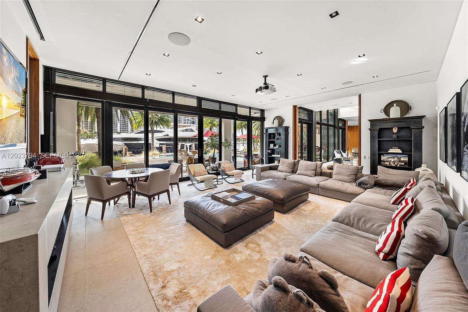 This expansive, open-concept living area features a large, neutral-toned sectional sofa paired with two leather ottomans, creating a comfortable space for relaxation. The room is bathed in natural light from floor-to-ceiling glass doors that open to an outdoor patio, while a dark, built-in shelving unit adds a sophisticated contrast to the bright walls. A dining area with a round table and modern chairs sits adjacent to the living space, and a projector mounted on the ceiling suggests a dedicated media setup.