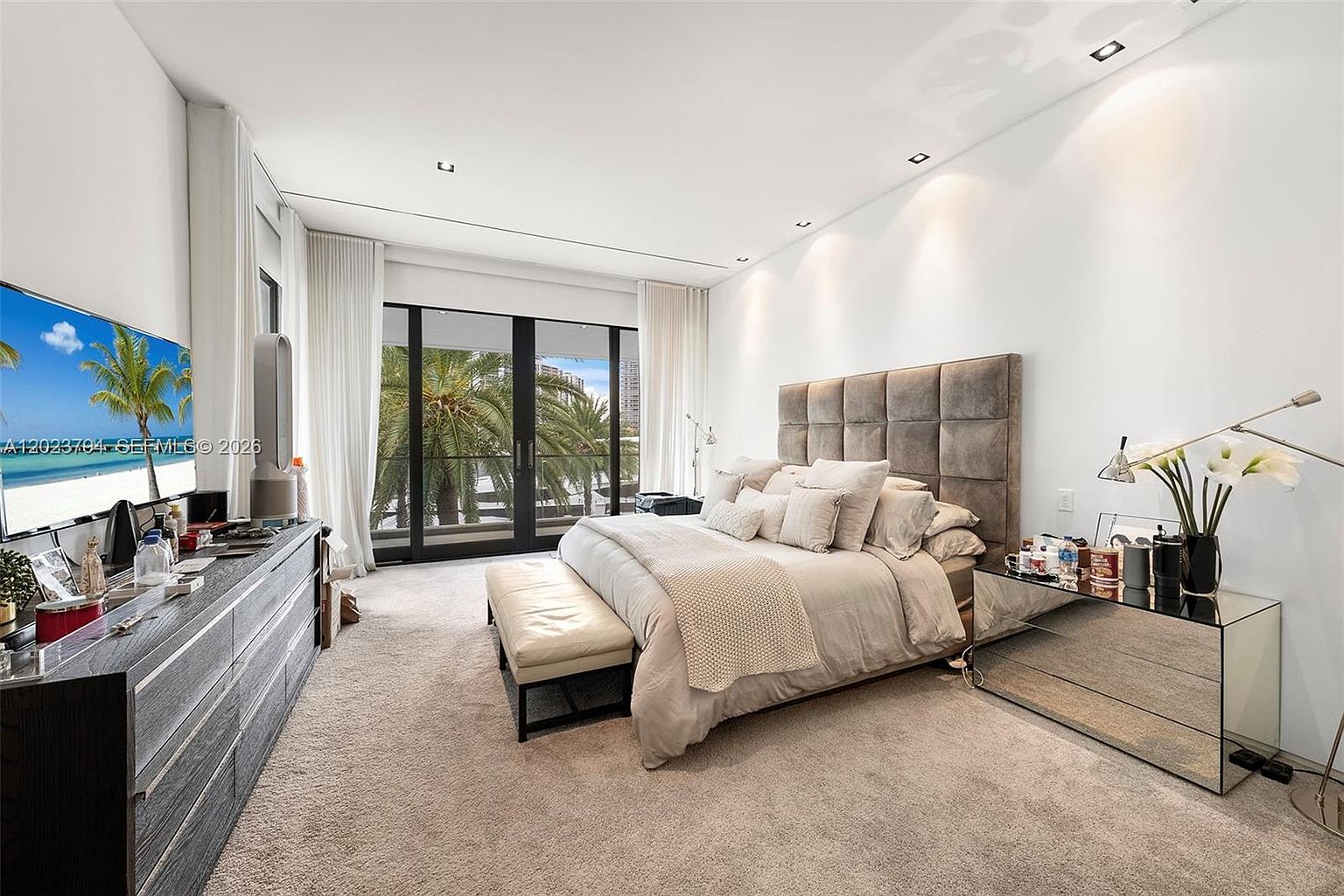 This spacious primary bedroom features a large, tufted upholstered headboard as the focal point, complemented by a neutral color palette and plush carpeting. A mirrored nightstand adds a touch of elegance, while floor-to-ceiling glass doors provide access to a balcony with lush palm tree views. The room is well-lit with recessed ceiling lights and a sleek, modern aesthetic.