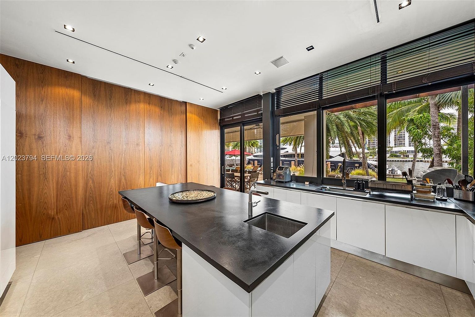 This modern kitchen features a large central island with a dark countertop and integrated sink, paired with three bar stools. The space is defined by extensive wood-paneled cabinetry on the left and a sleek white lower cabinet run along the window wall, which offers views of a waterfront area. Large windows with dark frames allow natural light to fill the room, creating a bright and contemporary atmosphere.