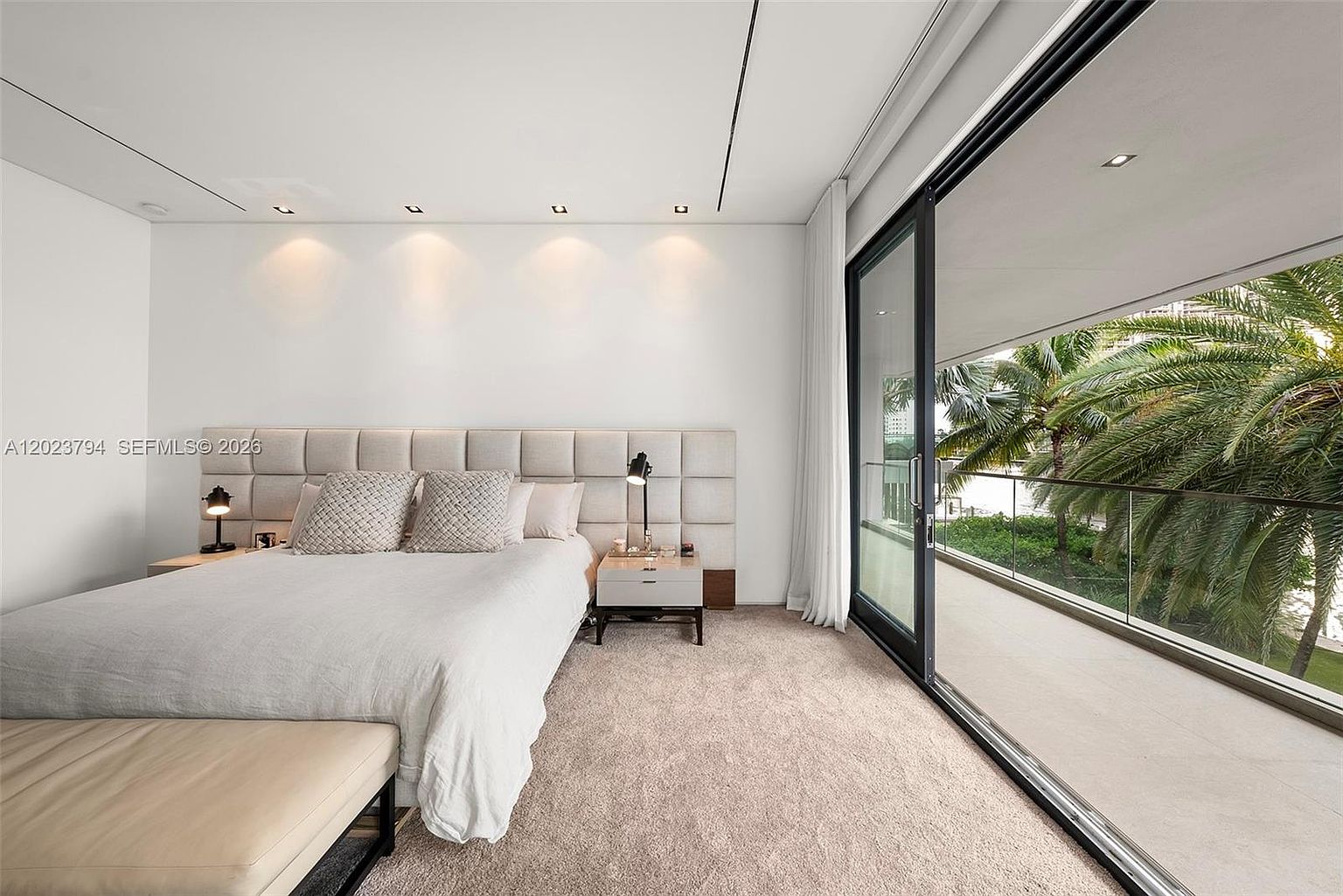 This primary bedroom features a contemporary design with a large, upholstered headboard wall and neutral-toned bedding. A wall of floor-to-ceiling glass sliding doors provides abundant natural light and direct access to a private balcony overlooking lush palm trees and water. The room is finished with soft carpeting and recessed lighting, creating a serene and upscale atmosphere.