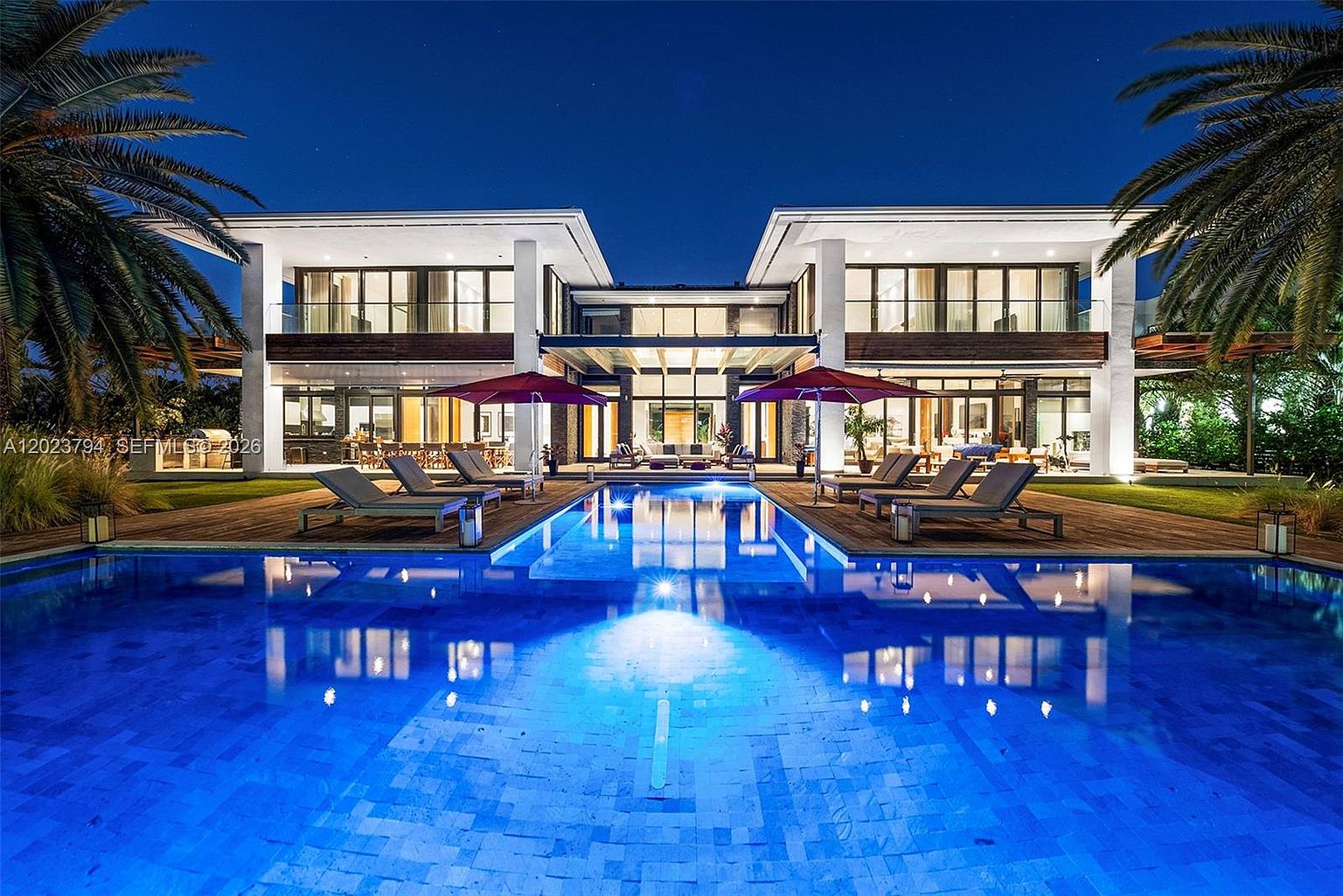 This stunning contemporary estate is captured at twilight, showcasing a symmetrical architectural design with a large, illuminated swimming pool as the central focal point. The residence features expansive glass walls and a wooden deck lined with lounge chairs and red umbrellas, creating a luxurious resort-like atmosphere. The scene is framed by lush palm trees, emphasizing the property's sophisticated and tranquil outdoor living space.