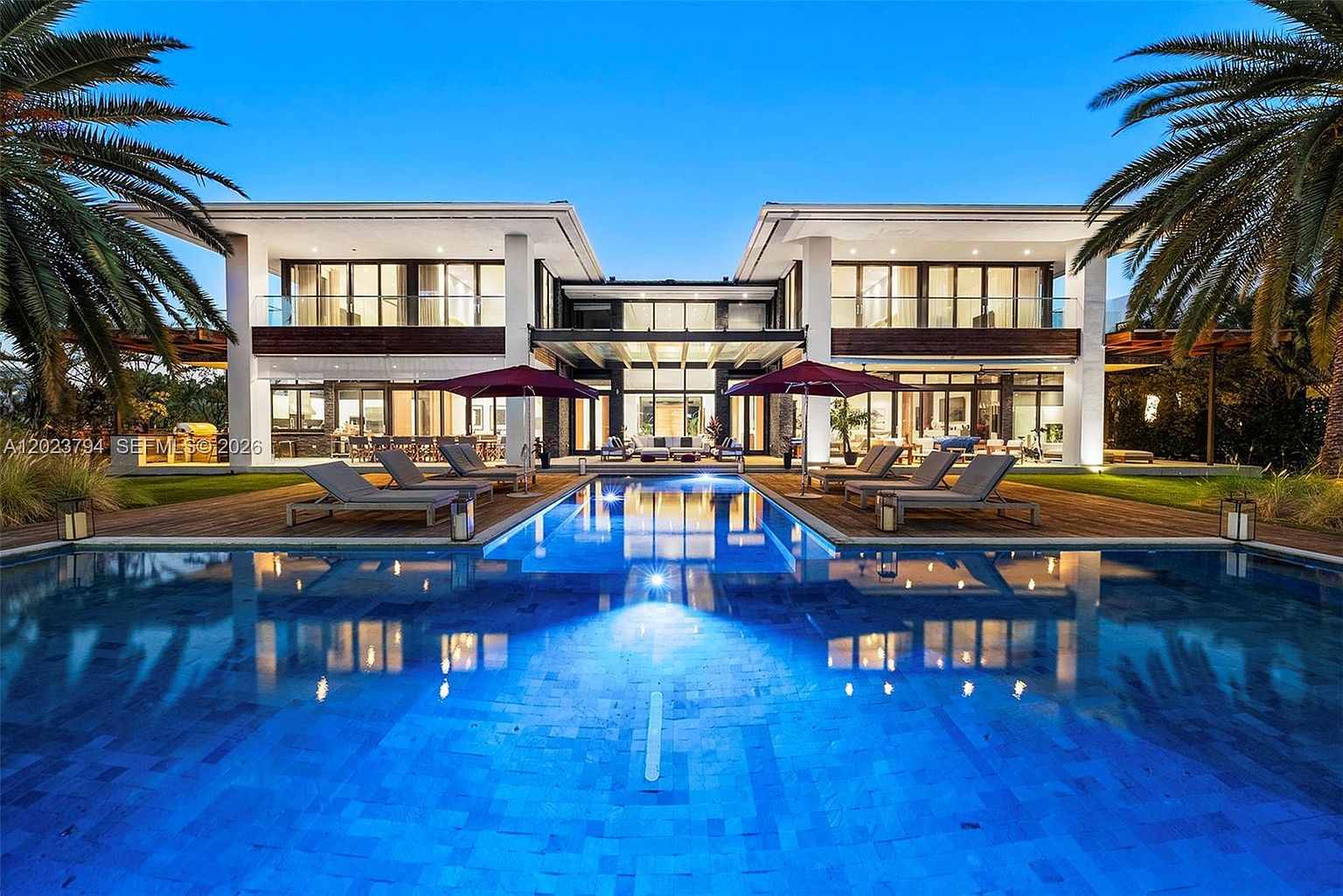 This stunning contemporary estate is captured from a low-angle perspective across a large, illuminated swimming pool, emphasizing the symmetry and modern architectural lines of the residence. The two-story structure features expansive glass walls, dark wood accents, and a central covered patio area flanked by two large red umbrellas. The scene is set at twilight, with the warm interior lighting contrasting beautifully against the deep blue sky and the reflective surface of the pool.