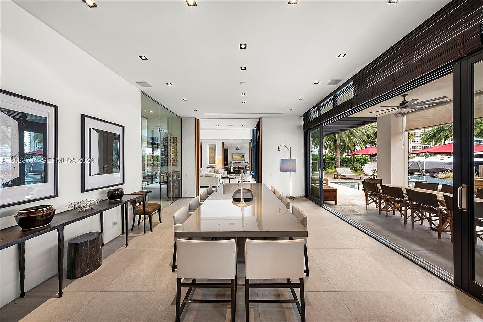 This sophisticated dining area features a long, polished table surrounded by modern, light-toned chairs, creating an elegant space for formal gatherings. The room seamlessly transitions to an outdoor patio through expansive sliding glass doors, which provide a clear view of the pool and lush landscaping. The interior is characterized by a minimalist aesthetic, neutral color palette, and recessed lighting, emphasizing a bright and airy atmosphere.