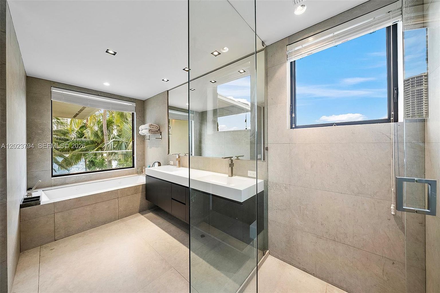 This modern primary bathroom features a sleek, dark-toned vanity with dual sinks and a large, frameless mirror. A deep soaking tub is positioned beneath a large window that offers a view of lush palm trees and water, while a glass-enclosed shower stall sits to the right. The space is finished with neutral, large-format stone tiles on the walls and floors, creating a clean and contemporary aesthetic.