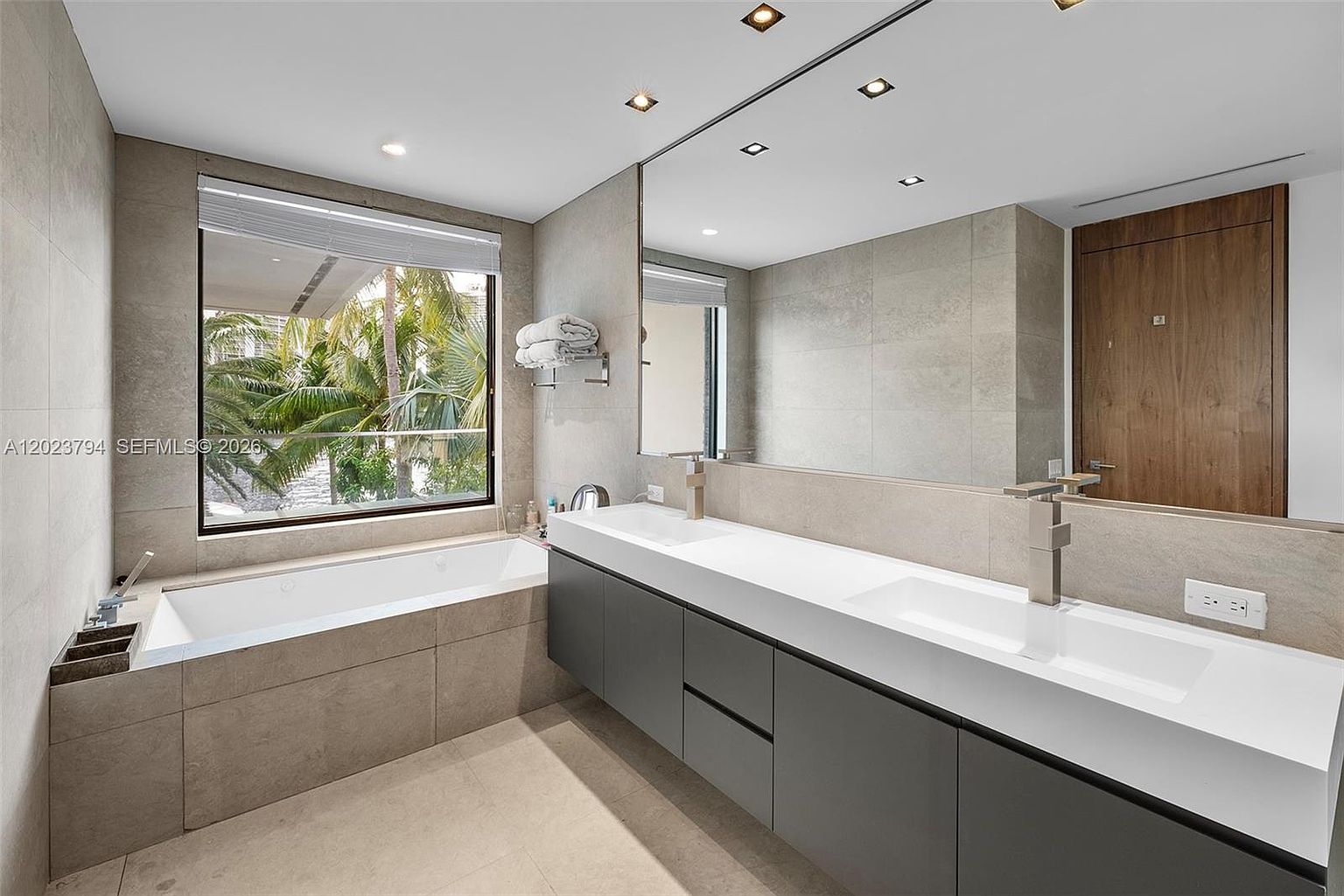 This modern primary bathroom features a sleek, floating double vanity with a long white countertop and dark grey cabinetry. A deep soaking tub is positioned beneath a large window that offers a view of lush greenery outside, while a large wall-mounted mirror reflects the space. The room is finished with neutral-toned wall and floor tiles, creating a clean and contemporary aesthetic.