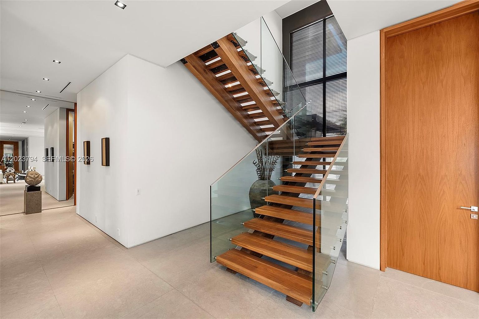 This modern entryway features a striking floating staircase with light wood treads and a clear glass railing, creating an open and airy aesthetic. The space is characterized by minimalist white walls, large-format light-toned floor tiles, and a tall wooden door that adds warmth to the clean design. A hallway extends to the left, showcasing a sculptural art piece and recessed lighting, while a large window at the top of the stairs allows natural light to fill the foyer.