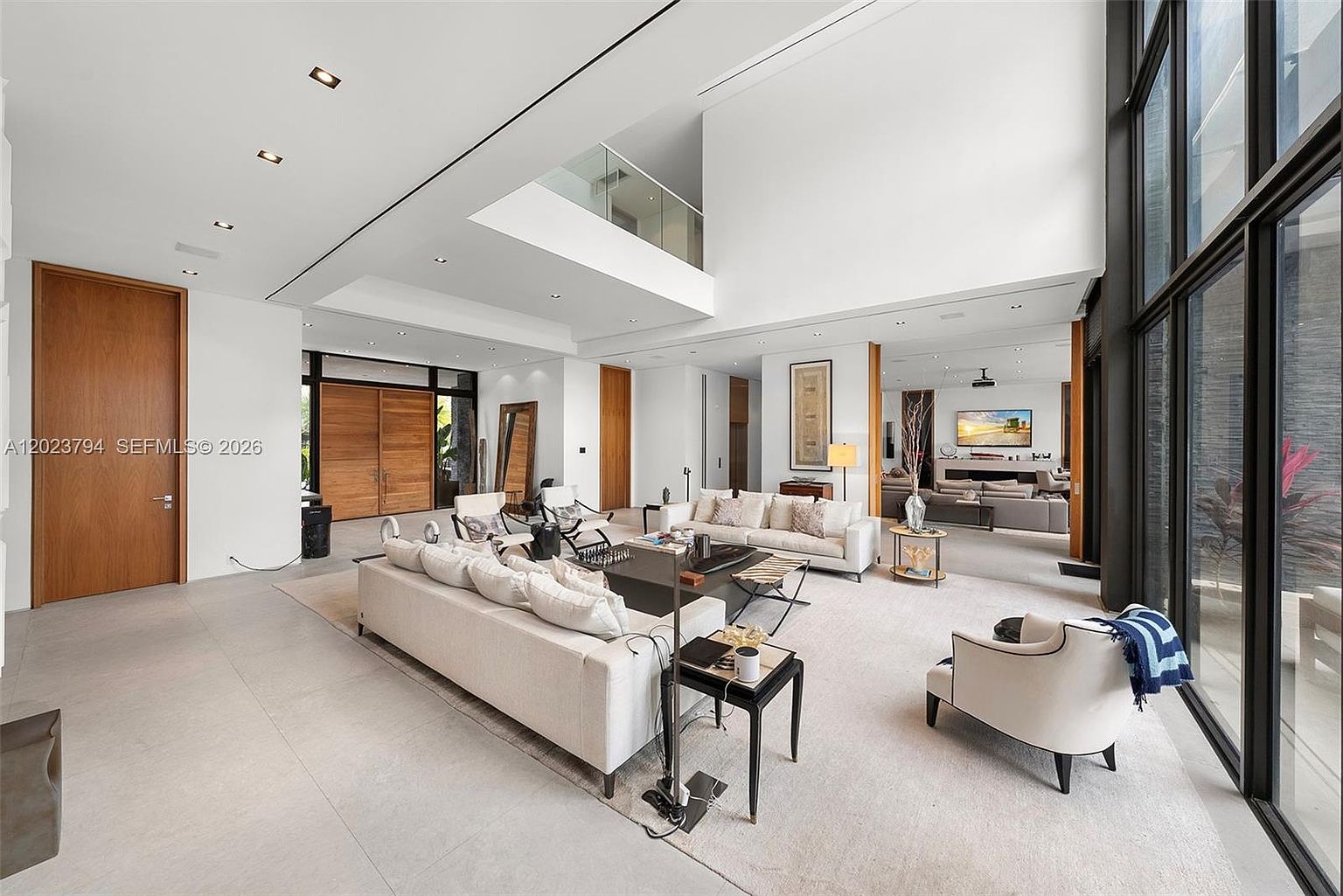 This expansive, double-height living room features a bright, open-concept design with neutral-toned furnishings and large windows that provide abundant natural light. The space includes a primary seating area with a large sofa and armchairs, leading into a secondary lounge area visible in the background. The minimalist aesthetic is complemented by clean lines, light-colored flooring, and a mezzanine level overlooking the main living space.