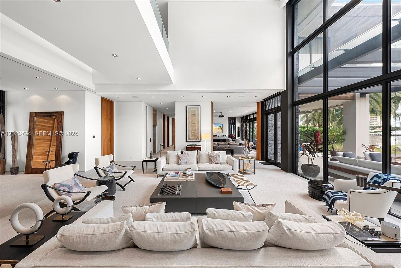 This expansive, double-height living room features a sophisticated open-concept layout with neutral-toned furnishings and a large, dark-toned central coffee table. The space is defined by massive floor-to-ceiling windows that provide abundant natural light and a seamless connection to the outdoor patio area. The minimalist aesthetic is complemented by high ceilings, clean lines, and a neutral color palette, creating an airy and luxurious atmosphere.