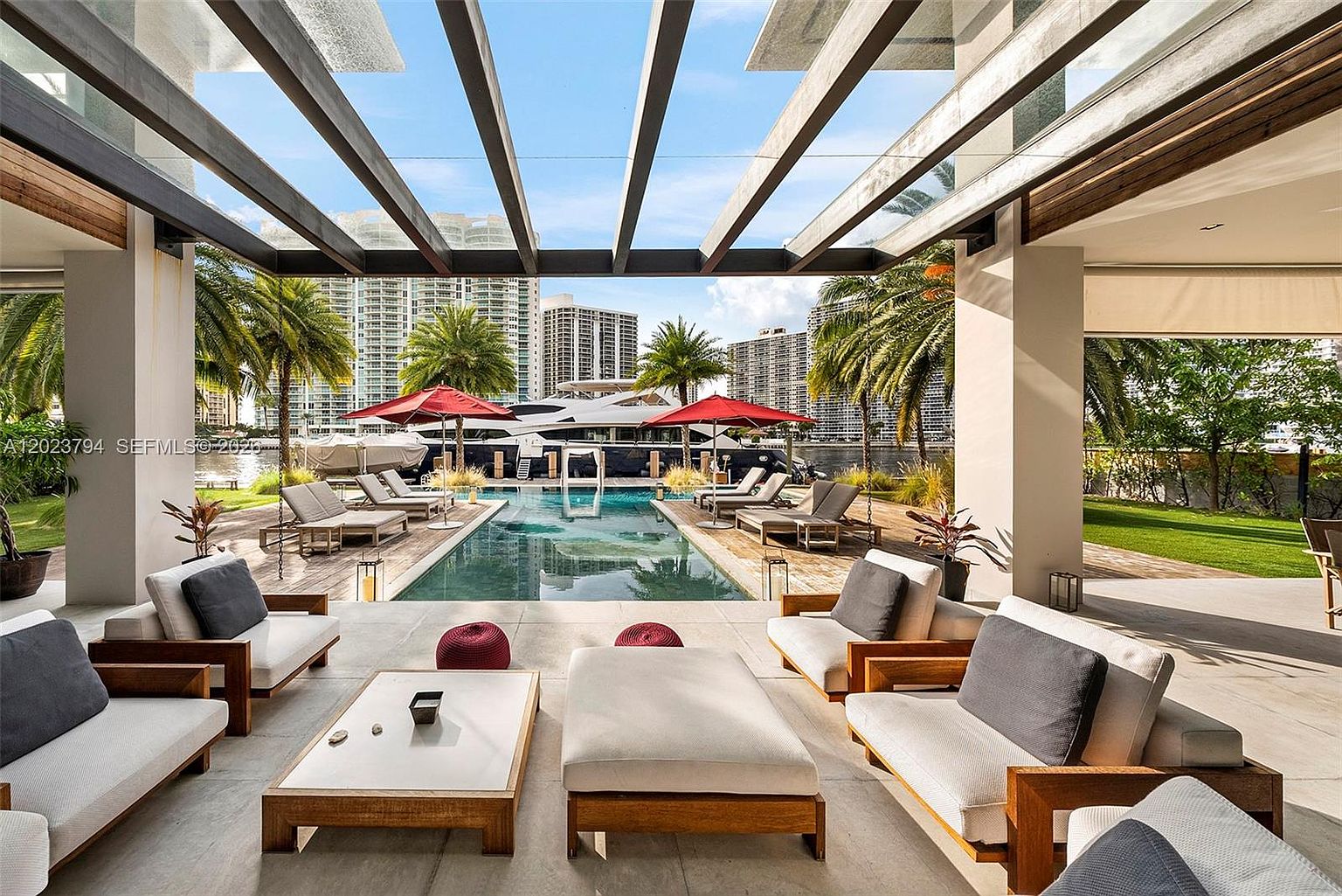 This luxurious covered patio offers a seamless transition to a private swimming pool and waterfront view. The space is furnished with modern, light-wood outdoor seating and a central coffee table, all sheltered by a sleek, open-beam pergola structure. Beyond the pool, a large yacht is docked, framed by lush palm trees and high-rise city buildings in the background.