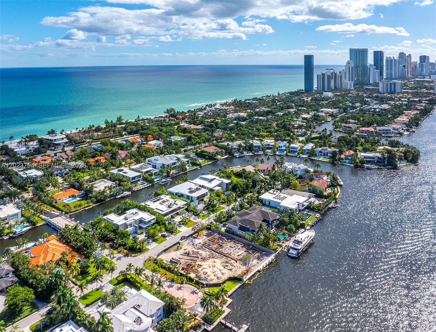 This high-altitude aerial view captures a luxurious waterfront neighborhood in a coastal city, showcasing a network of canals lined with modern, high-end residences. The scene features a prominent construction site in the foreground, a large yacht docked along a private seawall, and a sprawling cityscape with skyscrapers in the distance. The bright, sunny atmosphere highlights the turquoise ocean waters and the lush tropical landscaping throughout the residential area.