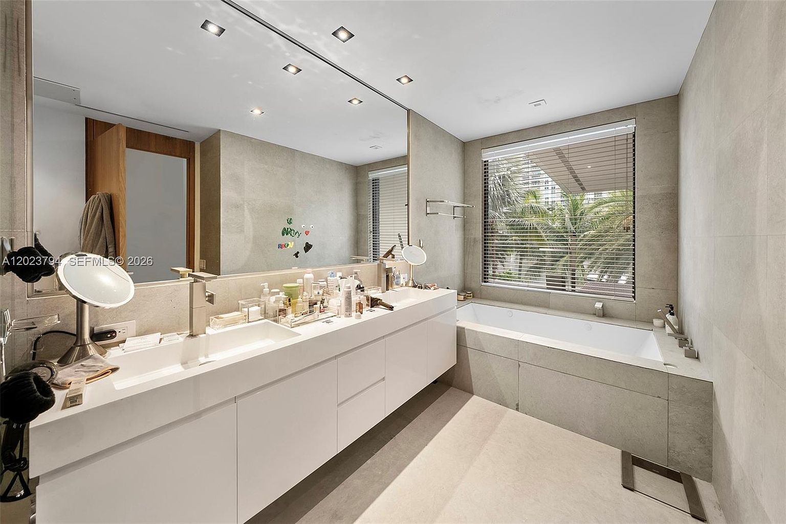 This primary bathroom features a long, white dual-sink vanity with ample counter space and a large, wall-to-wall mirror that enhances the room's brightness. To the right, a deep soaking tub is positioned beneath a large window covered by horizontal blinds, offering a view of palm trees outside. The space is finished with neutral-toned wall tiles and recessed ceiling lighting, creating a clean and functional atmosphere.