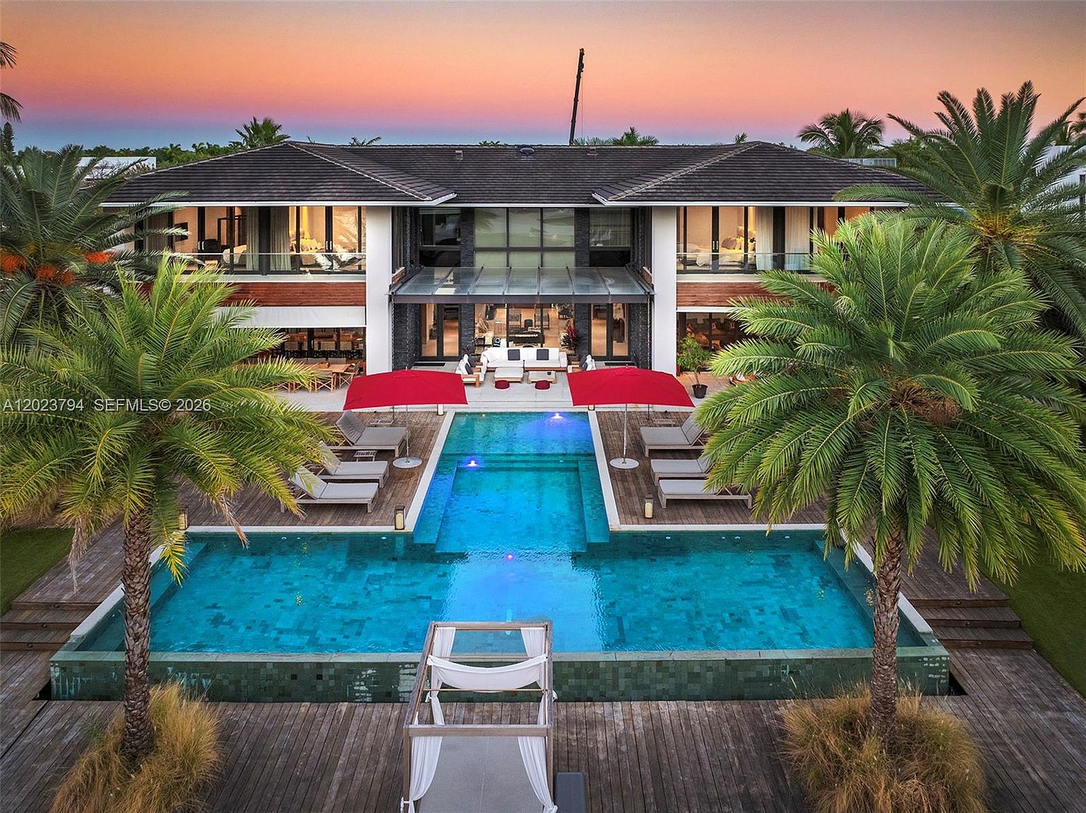 This stunning aerial perspective captures the rear elevation of a modern luxury residence at sunset, showcasing a symmetrical architectural design with expansive glass walls. The property features a multi-tiered swimming pool with a central lounge area, flanked by wooden decking, sun loungers, and two vibrant red umbrellas. Lush palm trees frame the scene, enhancing the tropical resort-like atmosphere of this high-end estate.