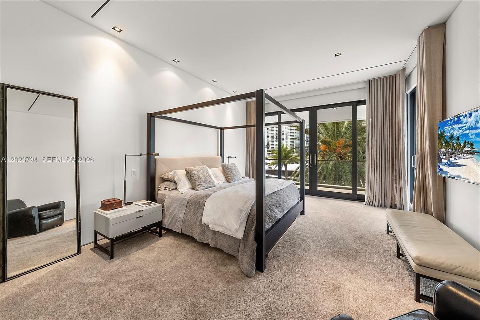 This spacious primary bedroom features a prominent four-poster bed as the central focal point, complemented by a sleek bedside table and a large floor-standing mirror. The room is bathed in natural light from expansive glass doors that lead to an outdoor area, while a long bench sits at the foot of the bed. The neutral color palette and clean lines create a sophisticated, modern atmosphere suitable for a luxury residence.