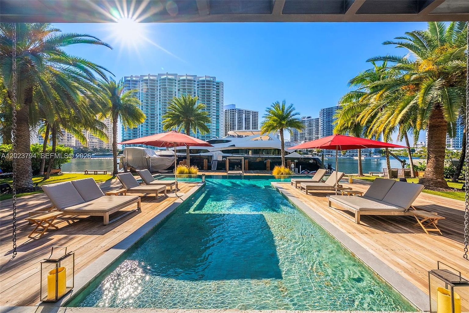 This luxurious waterfront pool area features a rectangular swimming pool surrounded by a light-toned wooden deck and multiple lounge chairs. Two red umbrellas provide shade, while lush palm trees frame the view of a large yacht docked in the canal against a backdrop of high-rise city buildings. The scene is captured from a low, centered perspective, emphasizing the serene atmosphere and the seamless transition between the private outdoor living space and the water.