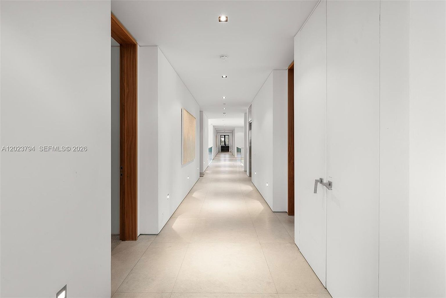 This long, minimalist hallway features bright white walls and large, light-toned floor tiles that create a clean, expansive aesthetic. The space is punctuated by warm-toned wood door frames and a piece of abstract art on the left wall, leading the eye toward a distant doorway at the end of the corridor. Recessed ceiling lighting provides a soft, uniform glow, emphasizing the sleek and modern architectural design of the home.