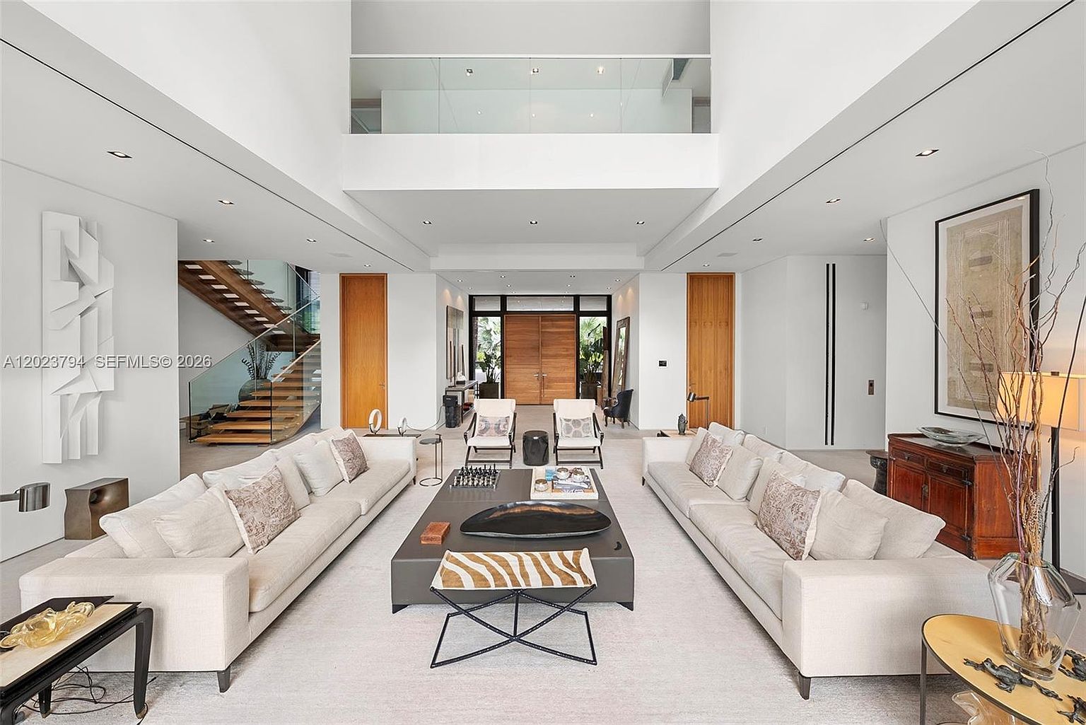 This expansive, double-height living room features a symmetrical layout with two large, light-colored sofas facing a central coffee table. The space is defined by its soaring ceilings, a glass-railed mezzanine overlooking the area, and a floating wooden staircase to the left. The minimalist aesthetic is complemented by neutral tones, contemporary art, and large wooden double doors at the far end, creating an airy and sophisticated atmosphere.