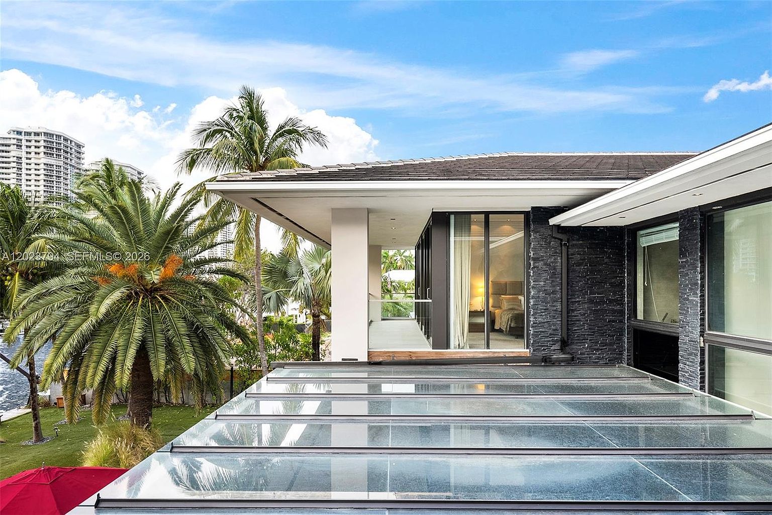 This elevated perspective captures a modern outdoor terrace featuring a glass-paneled roof structure that provides a sleek, contemporary aesthetic. The space connects to an interior room visible through large glass doors, while a lush palm tree and tropical foliage frame the view of the surrounding neighborhood. The combination of dark stone wall cladding and clean architectural lines creates a sophisticated, resort-like atmosphere.