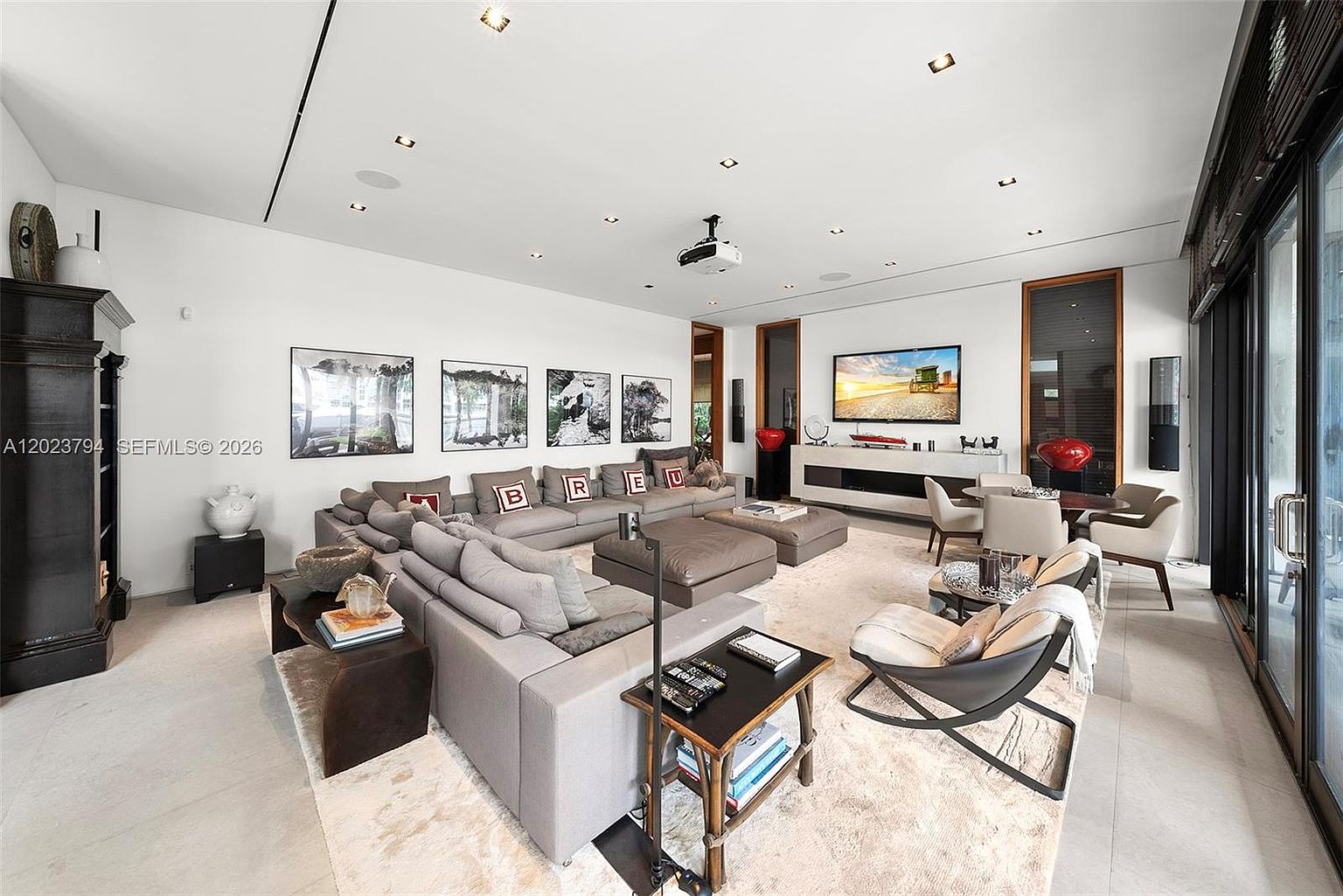 This spacious, contemporary living room features a large, neutral-toned sectional sofa adorned with decorative pillows spelling out 'BREU'. The room is anchored by a large area rug over light-colored flooring, with a low-profile media console beneath a wall-mounted television. Large glass sliding doors on the right provide ample natural light, while a projector mounted on the ceiling suggests a dedicated home theater setup.