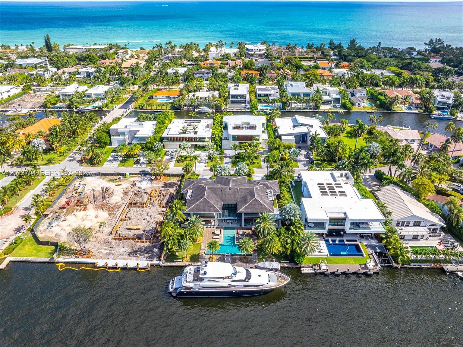This high-angle aerial perspective captures a luxurious waterfront estate in a dense residential neighborhood, featuring a large yacht docked at the private seawall. The property includes a multi-level residence with a swimming pool, adjacent to a construction site and a neighboring modern home with a sleek, flat-roof design. The scene showcases the expansive canal system leading toward the open ocean in the background, emphasizing the prime coastal location.
