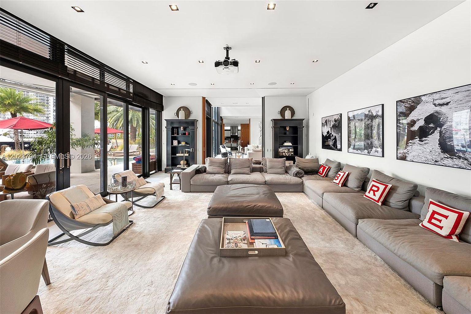This expansive, contemporary living room features a massive modular sectional sofa adorned with decorative pillows, centered around two large leather ottomans. The space is bathed in natural light from floor-to-ceiling glass sliding doors that open to a pool deck, while a projector mounted on the ceiling suggests a dedicated media area. The room is decorated with large-scale black and white photography and symmetrical dark shelving units, creating a sophisticated and airy atmosphere.