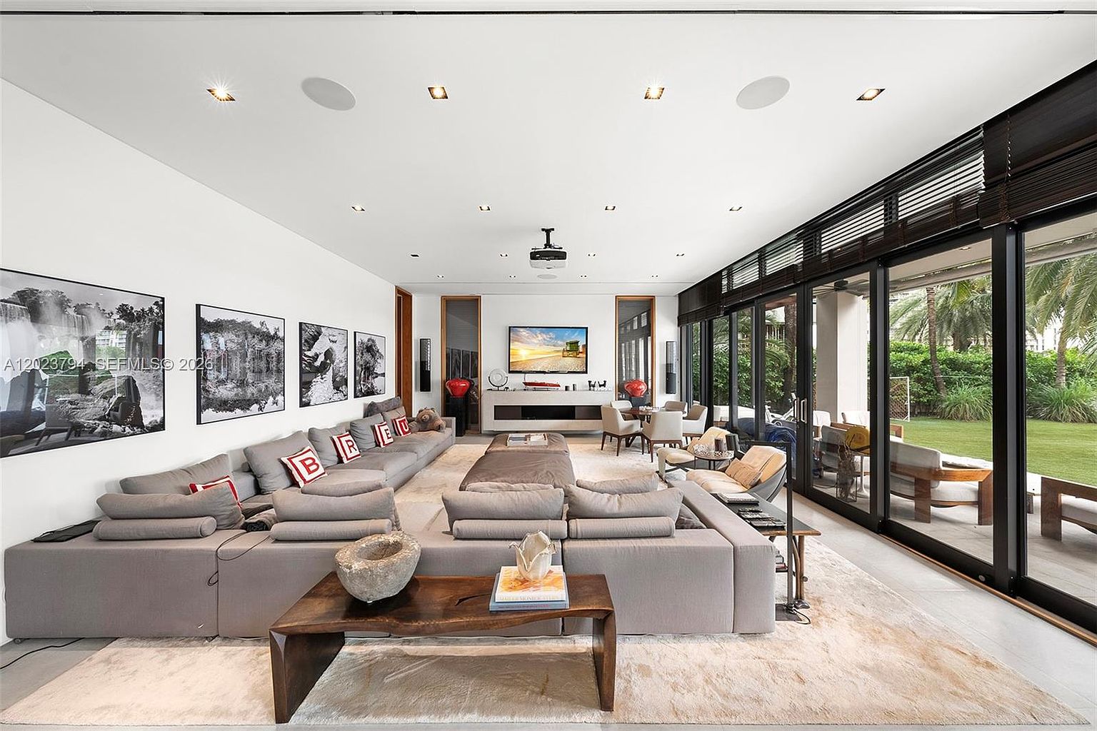 This expansive, contemporary living room features a massive, L-shaped sectional sofa arranged around a central coffee table, creating a comfortable lounge area. The space is defined by a long wall adorned with framed black-and-white photography and a wall of floor-to-ceiling glass sliding doors that provide a seamless view of the lush green backyard. A projector mounted on the ceiling and a television centered on the far wall highlight the room's function as a dedicated media and entertainment space.