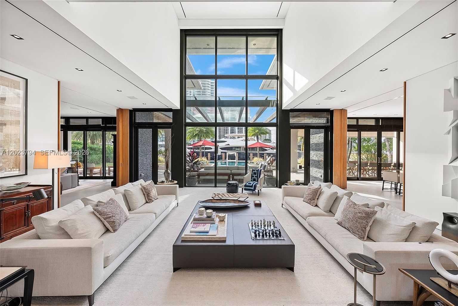 This expansive, high-ceilinged living room features a symmetrical layout with two large, light-colored sofas facing a central oversized ottoman coffee table. The space is anchored by a massive, multi-pane window wall that provides a clear view of the outdoor pool area and tropical landscaping. The room is designed with a clean, contemporary aesthetic, utilizing neutral tones and minimalist decor to emphasize the architectural scale and natural light.