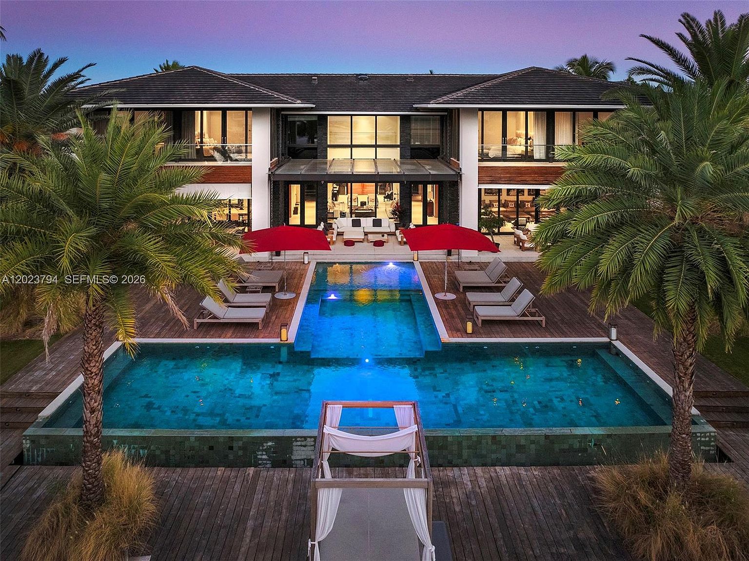 This stunning rear view of a luxury estate captures a symmetrical architectural design with a large, multi-level swimming pool as the central focal point. The property features a spacious wooden deck furnished with lounge chairs and red umbrellas, leading directly into the residence through large glass doors. The scene is set during twilight, with the warm interior lighting contrasting beautifully against the deep blue sky and the illuminated pool water.