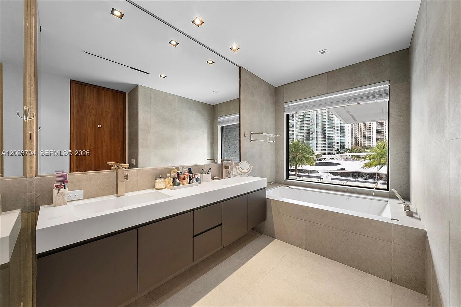 This modern primary bathroom features a long, dual-sink vanity with dark cabinetry and a large, wall-to-wall mirror that enhances the sense of space. To the right, a built-in soaking tub is positioned beneath a large window that provides natural light and views of the surrounding urban landscape. The room is finished with neutral-toned wall and floor tiles, creating a clean, contemporary, and spa-like atmosphere.