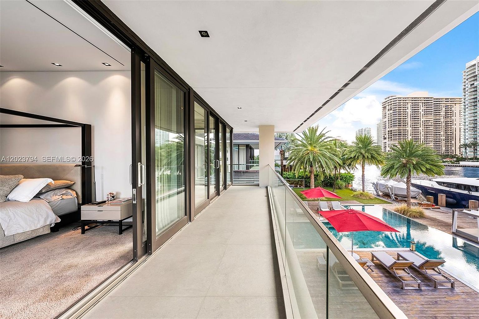 This image captures a spacious, modern balcony overlooking a private pool area and a waterway with docked boats. The balcony features large glass sliding doors that open into a bedroom, creating a seamless transition between indoor and outdoor living spaces. The scene is framed by a glass railing, offering unobstructed views of the lush landscaping and the high-rise buildings in the distance.