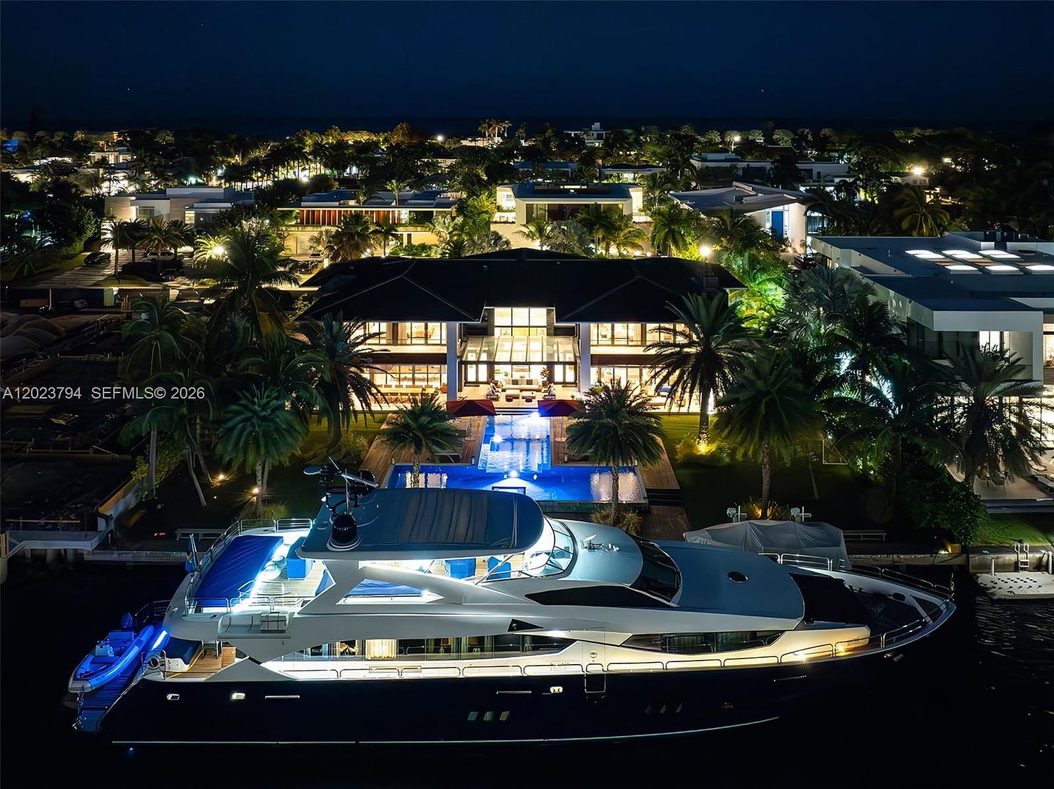 This stunning aerial night shot captures a luxurious waterfront estate featuring a large, multi-story modern residence illuminated against the dark sky. In the foreground, a sleek, dark-hulled yacht is docked along the canal, while the property itself boasts a glowing swimming pool and lush tropical landscaping. The perspective provides a cinematic overview of the home's architectural scale and its prime position on the water.
