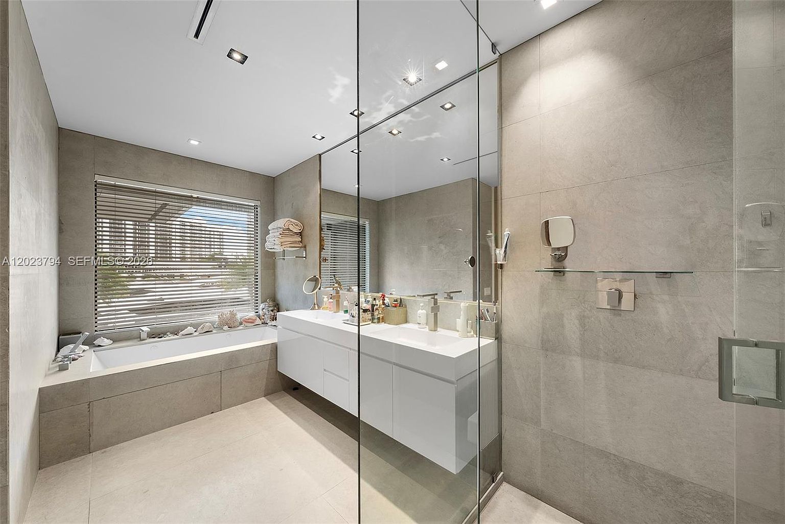 This modern primary bathroom features a sleek, floating double vanity with integrated sinks and a large, wall-to-wall mirror that enhances the sense of space. To the left, a deep soaking tub is positioned beneath a window with horizontal blinds, while a glass-enclosed shower stall is visible on the right. The room is finished with neutral, large-format stone tiles on both the walls and floors, creating a clean and minimalist aesthetic.