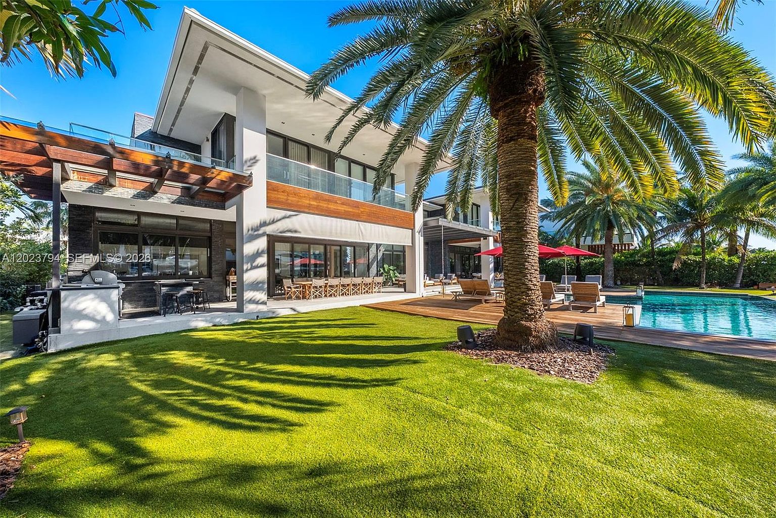 This luxurious modern residence features a sprawling rear exterior with a large, manicured lawn and a prominent palm tree as a focal point. The home showcases a contemporary design with expansive glass sliding doors, a wooden-accented second-story balcony, and a covered outdoor kitchen area. A wooden deck surrounds a sparkling swimming pool, creating an inviting space for outdoor entertaining and relaxation.