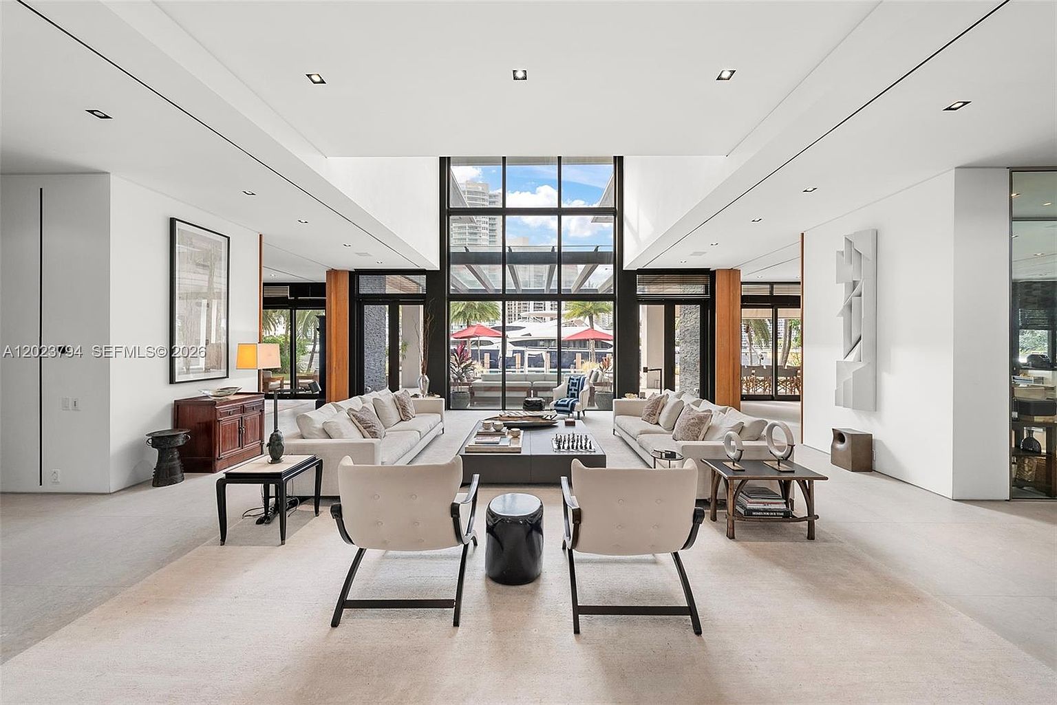 This expansive, high-ceilinged living room features a symmetrical layout with two large sofas facing a central coffee table and a pair of modern armchairs in the foreground. The space is anchored by a massive, multi-pane window wall that provides a clear view of the outdoor patio and waterfront beyond. The minimalist aesthetic is enhanced by neutral tones, recessed lighting, and clean architectural lines, creating an airy and sophisticated atmosphere.