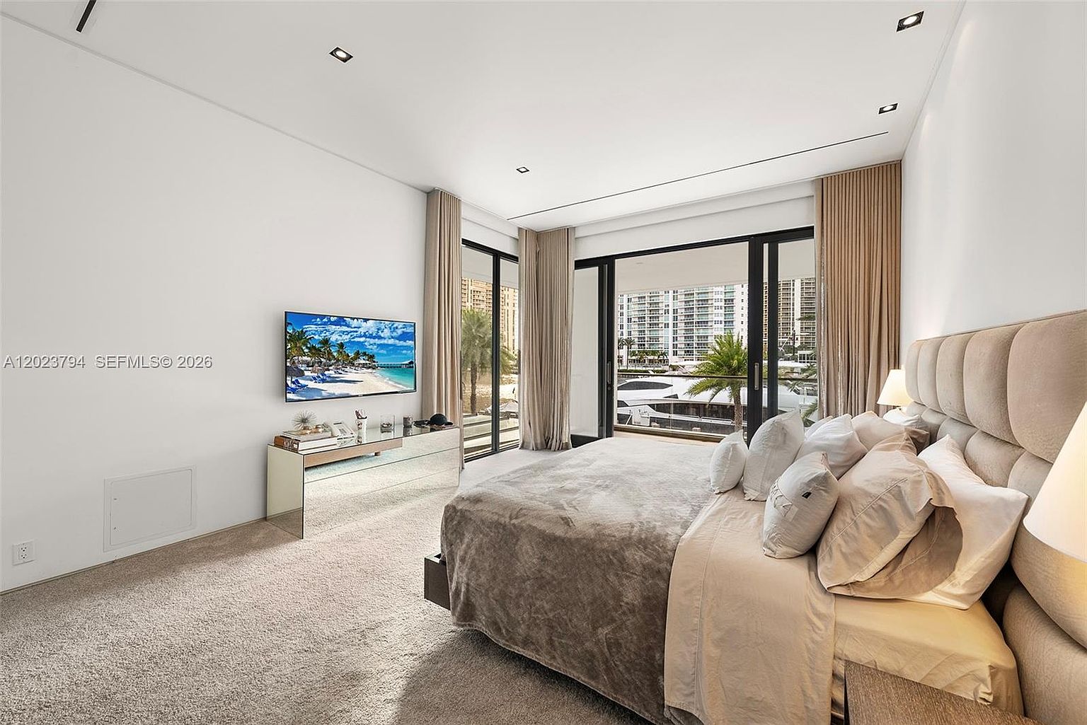 This spacious primary bedroom features a large, tufted upholstered headboard and a neutral color palette that creates a serene atmosphere. The room is highlighted by floor-to-ceiling windows that provide ample natural light and a view of the marina and surrounding high-rise buildings. A mirrored console table sits beneath a wall-mounted television, adding a touch of modern elegance to the space.