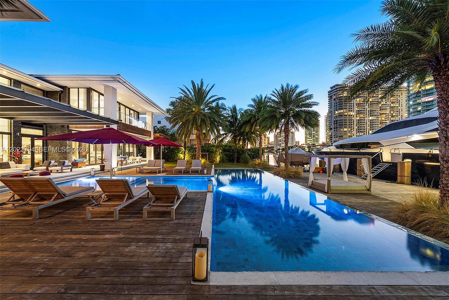This luxurious waterfront outdoor space features a long, rectangular swimming pool set into a spacious wooden deck. The area is furnished with multiple lounge chairs, umbrellas, and a shaded cabana, all overlooking a canal with a large yacht docked nearby. The modern architecture of the home is visible in the background, complemented by palm trees and a clear blue sky, creating an upscale, resort-like atmosphere.