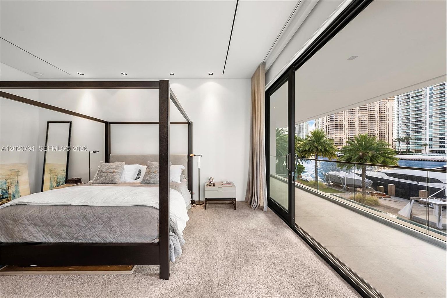 This primary bedroom features a dark wood four-poster bed as the central focal point, complemented by neutral bedding and a minimalist bedside table. Large floor-to-ceiling sliding glass doors open onto a private balcony, providing an expansive view of the waterfront and surrounding high-rise buildings. The room is finished with light-toned carpeting and recessed ceiling lighting, creating a serene and sophisticated atmosphere.
