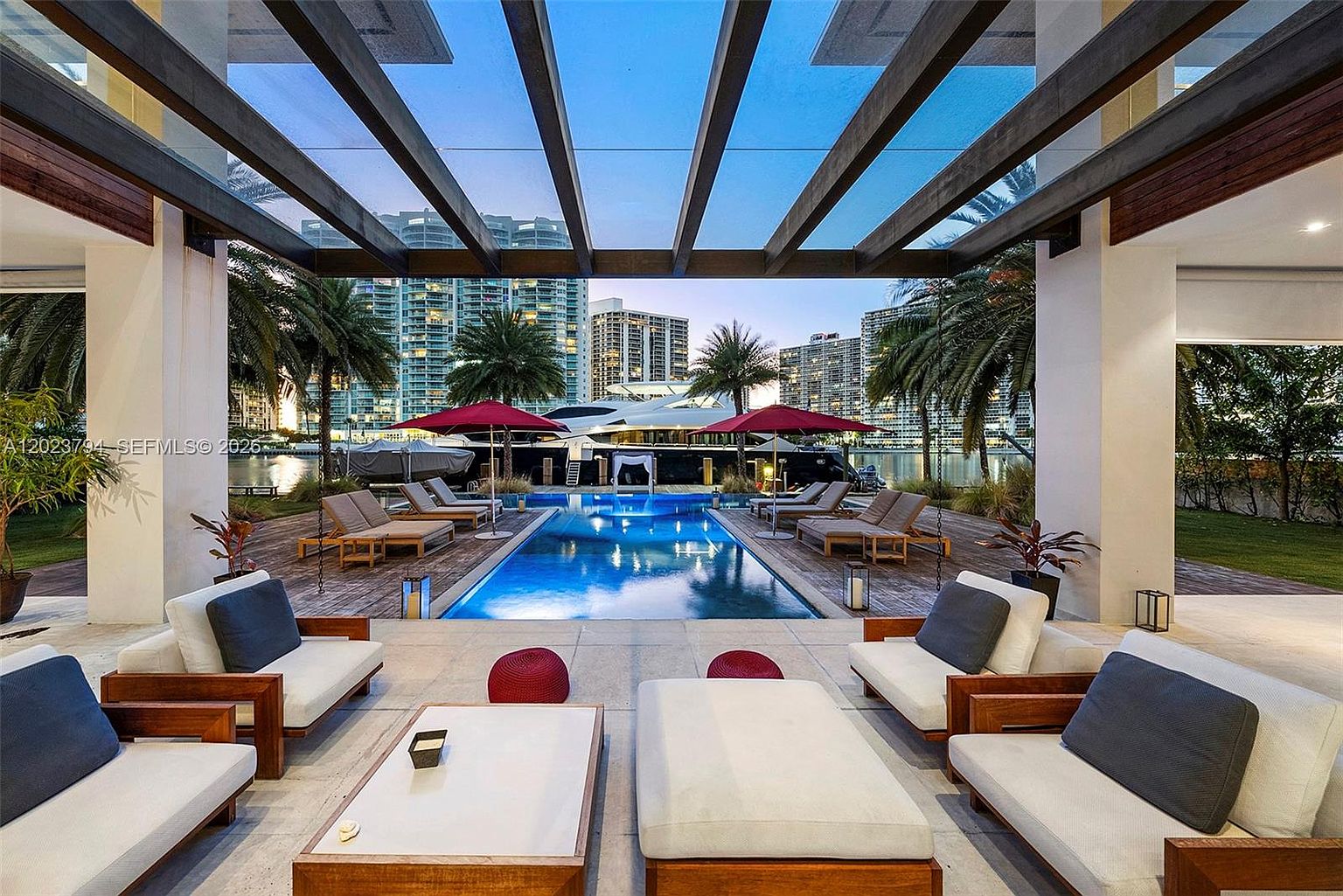 This luxurious outdoor living space features a covered patio area with comfortable lounge seating, leading directly to a rectangular swimming pool. The scene is framed by a wooden pergola structure, with a backdrop of palm trees, high-rise buildings, and a large yacht docked at the water's edge. The perspective is centered, capturing the seamless transition between the shaded lounge area and the sun-drenched pool deck at twilight.