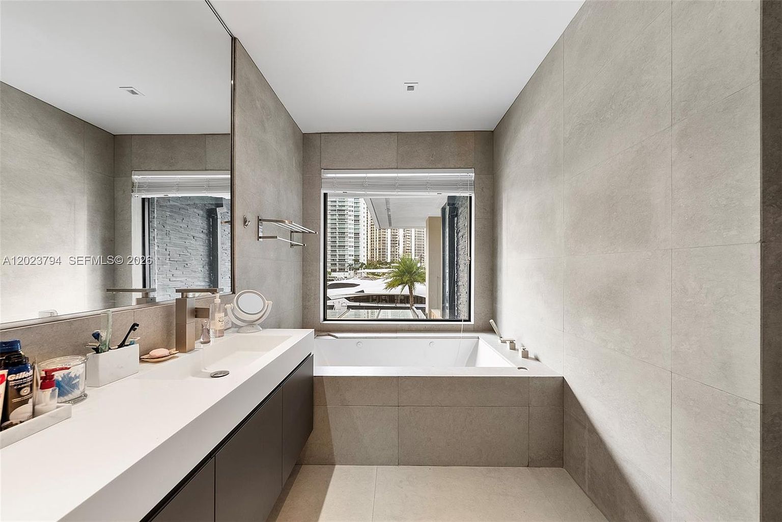 This modern primary bathroom features a sleek, minimalist design with large-format neutral-toned wall tiles and a dark vanity cabinet. A deep soaking tub is positioned directly beneath a large window that offers a view of the surrounding cityscape and marina. The space is bright and airy, highlighted by a wide mirror above the vanity and clean, contemporary fixtures.