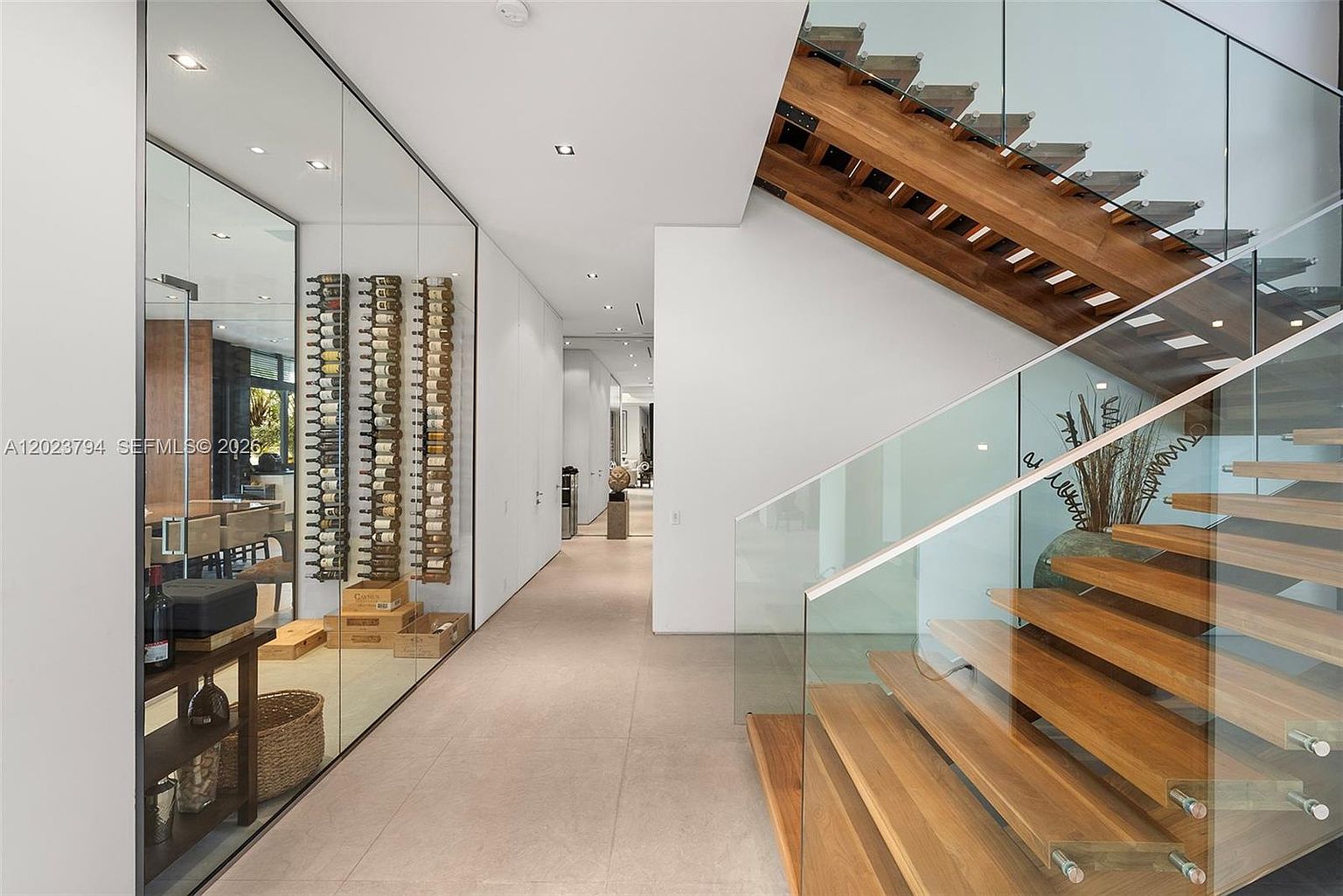 This modern interior features a striking floating staircase with light wood treads and a glass railing, creating an open and airy aesthetic. To the left, a glass-enclosed wine display adds a sophisticated architectural element, while a long, minimalist hallway leads toward the back of the home. The neutral color palette and clean lines emphasize a contemporary, high-end design.
