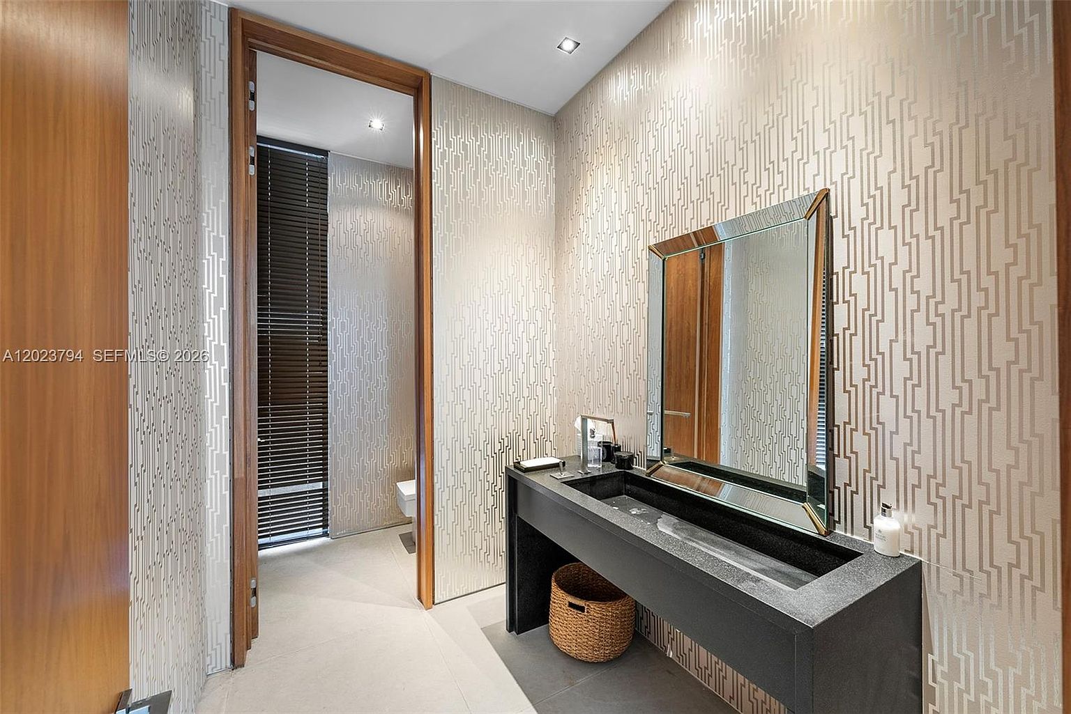 This modern powder room features a striking, dark stone trough-style sink set against a wall with intricate, geometric patterned wallpaper. A large, framed mirror hangs above the vanity, reflecting the sophisticated, neutral-toned space. A doorway leads to an adjacent area with dark blinds, adding depth and a sense of architectural flow to the room.