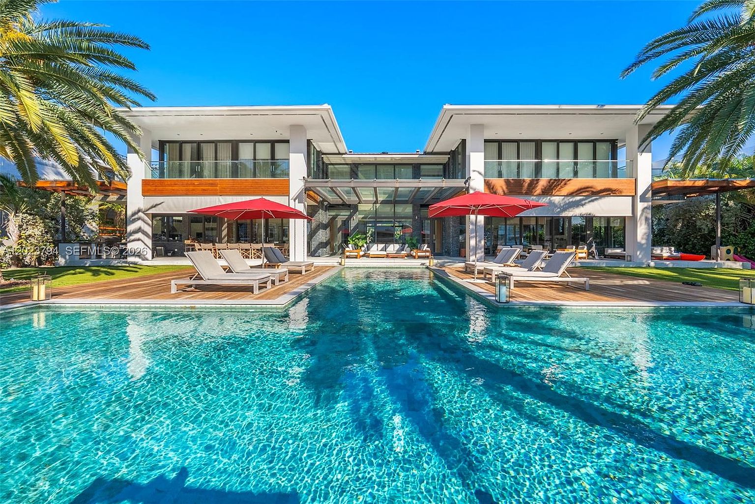 This stunning contemporary residence features a symmetrical architectural design with a large, rectangular swimming pool as its centerpiece. The expansive wooden deck is furnished with multiple lounge chairs and vibrant red umbrellas, creating a luxurious outdoor living space. Large glass panels on the rear facade provide a seamless transition between the indoor and outdoor areas, emphasizing a modern, resort-like aesthetic.