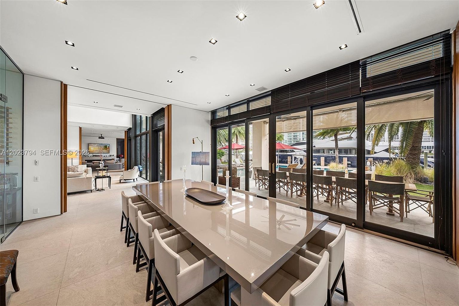 This spacious dining room features a large, glossy rectangular table surrounded by modern upholstered chairs, creating an elegant setting for formal meals. The room is defined by expansive floor-to-ceiling glass sliding doors that provide a seamless transition to the outdoor patio and pool area. A neutral color palette and recessed lighting enhance the bright, airy atmosphere, while a glimpse into the adjacent living area highlights the open-concept layout of the home.
