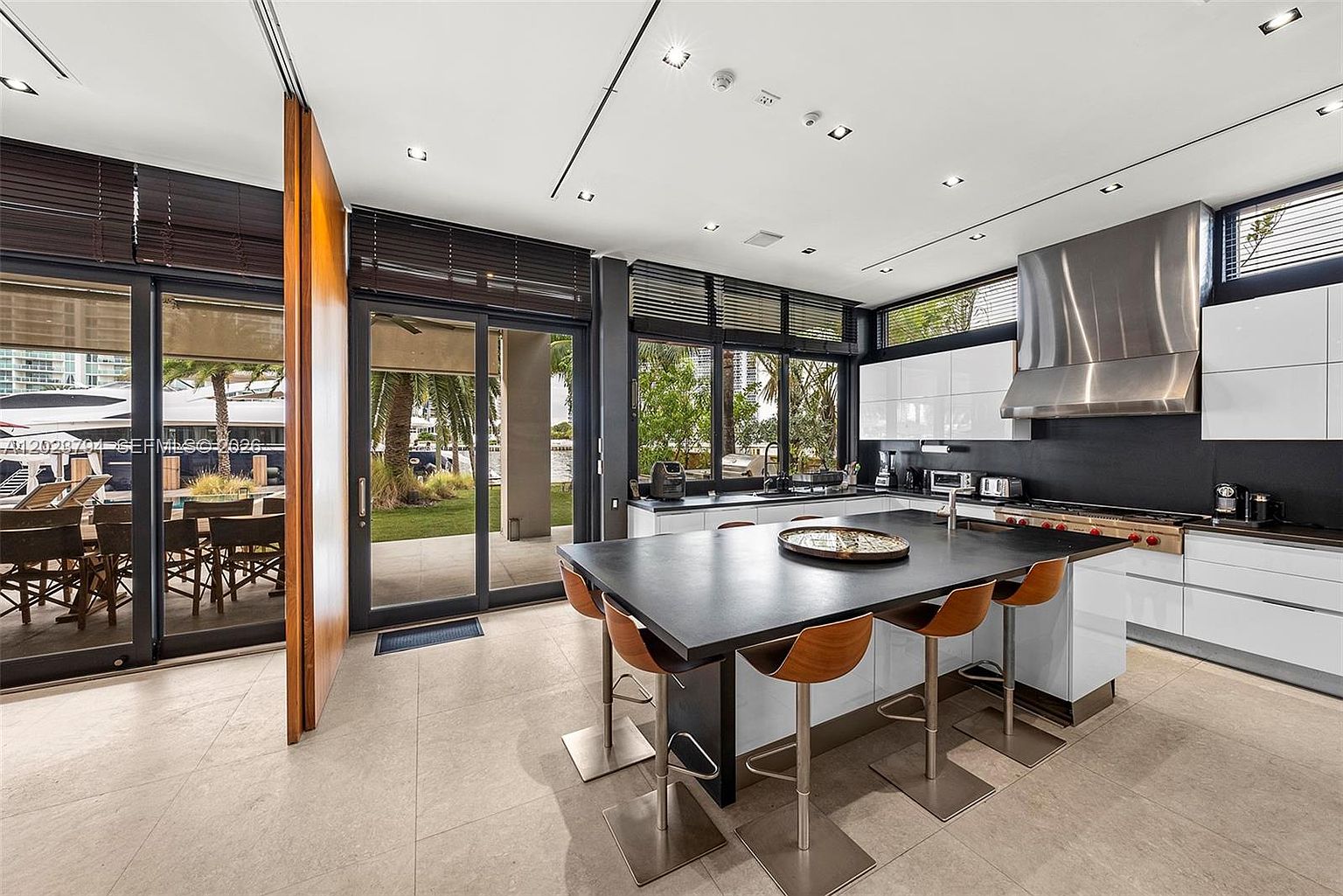 This modern kitchen features a large central island with a dark countertop and four wooden bar stools, serving as the focal point of the room. The space is equipped with white cabinetry, a stainless steel range hood, and professional-grade appliances, all set against a dark backsplash. Large sliding glass doors provide an expansive view of the outdoor patio and waterfront, creating a seamless transition between indoor and outdoor living areas.