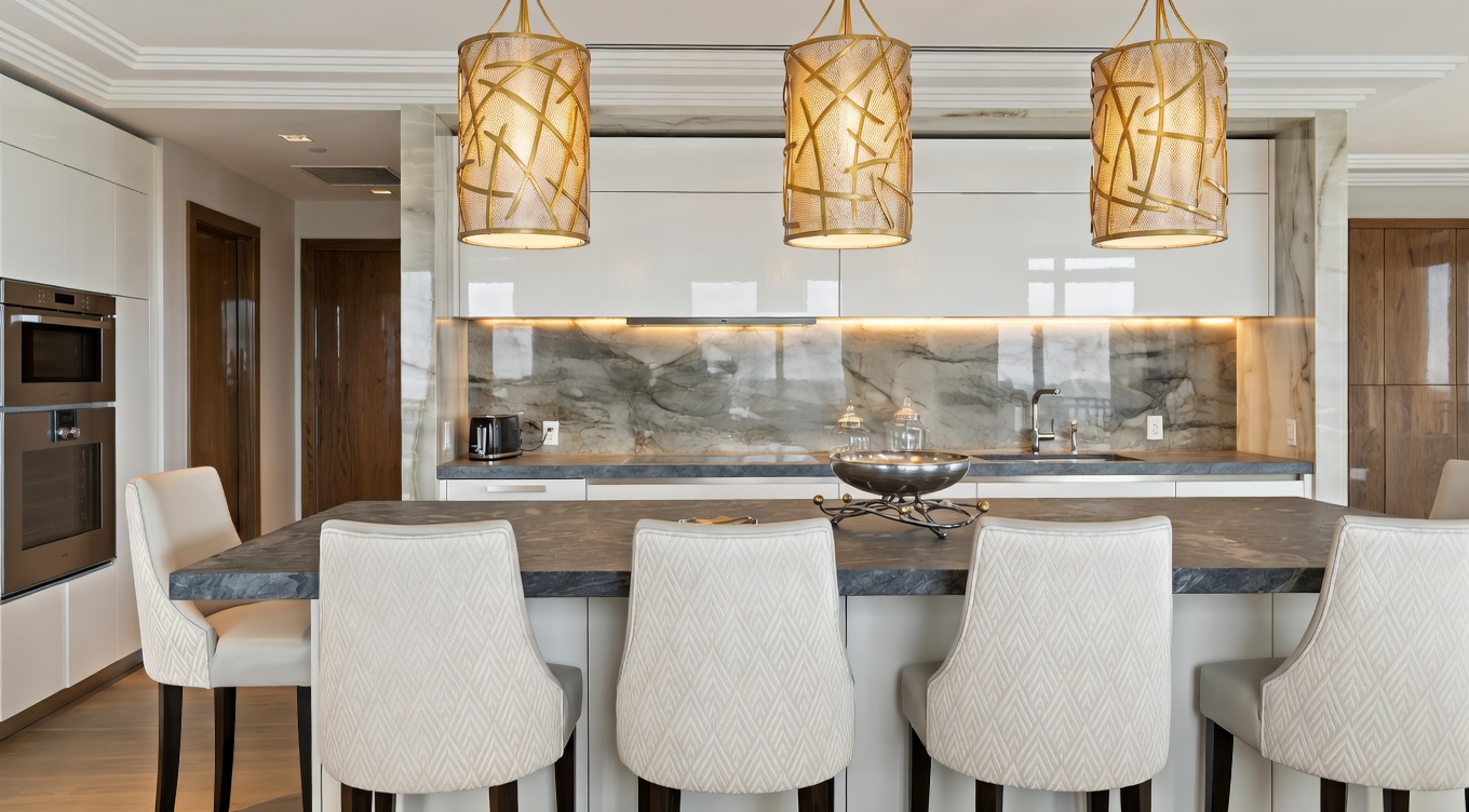 This sleek, modern kitchen features a large dark stone island with coordinated cream-colored high-back chairs, perfect for casual dining. The space is highlighted by a striking marble-effect backsplash and trio of textured gold pendant lights, complemented by minimalist white cabinetry and stainless steel appliances. The overall design conveys a sense of contemporary sophistication and high-end luxury.