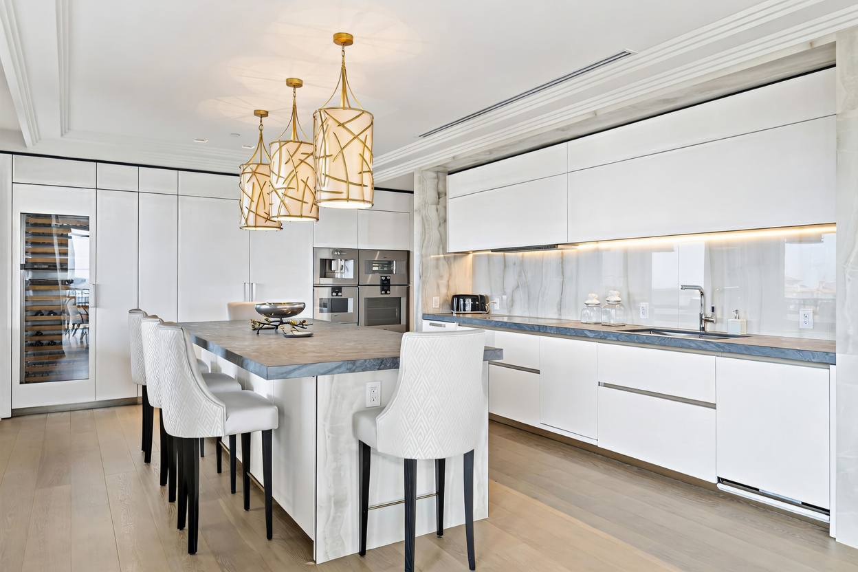 This ultra-modern, high-end kitchen features sleek white cabinetry, a large stone-topped island with integrated seating, and sophisticated gold-toned pendant lighting. The space is accented by a dramatic marble backsplash with integrated under-cabinet lighting and high-end built-in appliances, creating a luxurious and minimalist aesthetic. The perspective is a wide, eye-level shot that captures the seamless flow between the cooking area and the dining island.