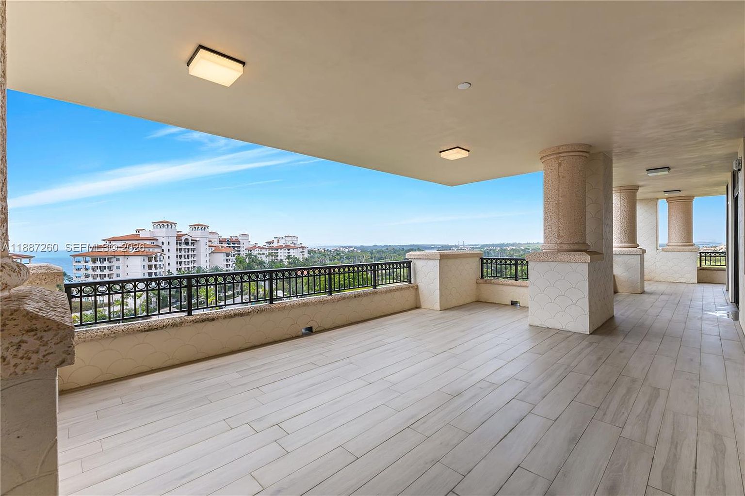 This expansive, covered balcony features light-toned wood-look tile flooring and sturdy, decorative columns that frame a stunning panoramic view of the surrounding landscape and distant water. The space is enclosed by a black metal railing, offering a secure yet open-air environment perfect for relaxation or entertaining. The neutral color palette and clean lines create a sophisticated, airy atmosphere that emphasizes the property's prime location.